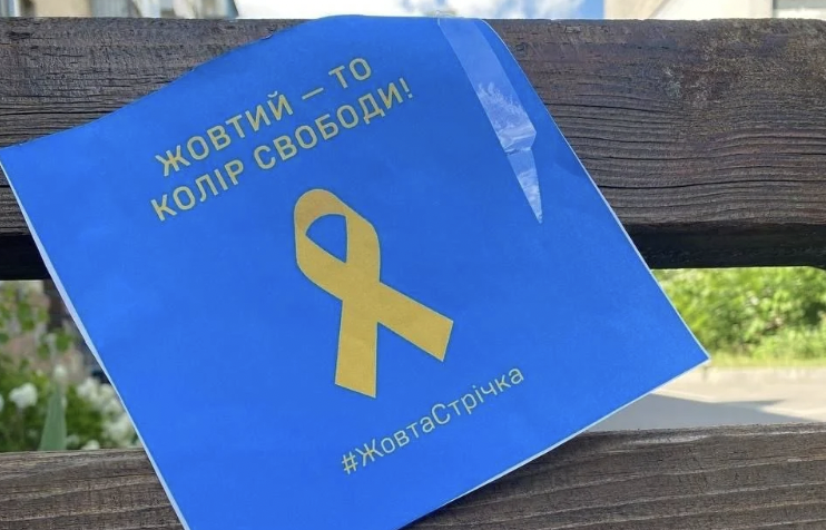 Синє оголошення на дерев'яній поверхні із жовтою стрічкою та текстом українською: "ЖОВТИЙ — ТО КОЛІР СВОБОДИ!" та хештегом "#ЖовтаСтрічка"; символ українського спротиву до Міжнародного дня руху опору