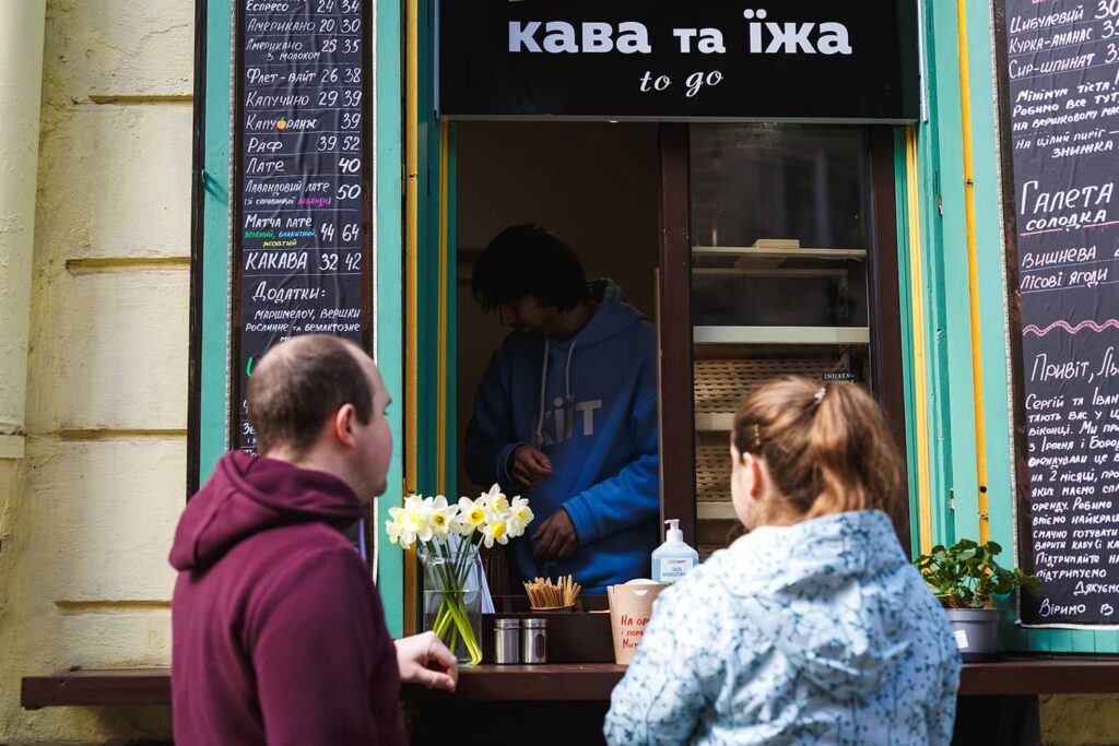 Вуличне віконце кав'ярні, всередині якого бариста готує замовлення. Поруч на стіні крейдяні дошки з написами асортименту, а перед віконцем стоять покупці.