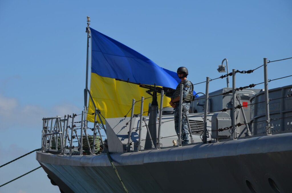Військовослужбовець ВМС ЗСУ у камуфляжній формі, касці та бронежилеті стоїть на палубі військового корабля біля кулемета.